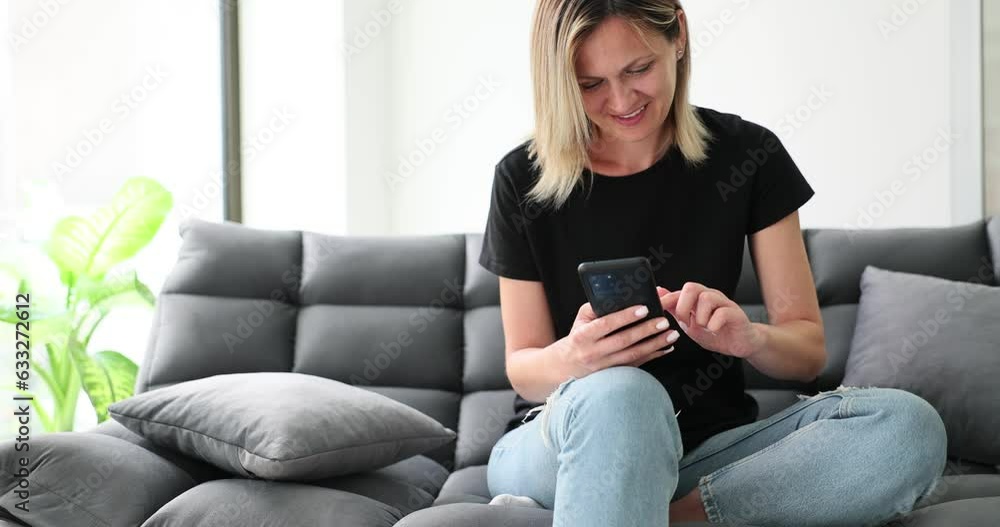 Smiling girl relaxing, sitting on sofa at home using mobile phone and chatting with friend. Happy young woman on sofa checking mobile app or social media Smiling girl relaxing, sitting on sofa at home using mobile phone and chatting with friend. Happy young woman on sofa checking mobile app or social media