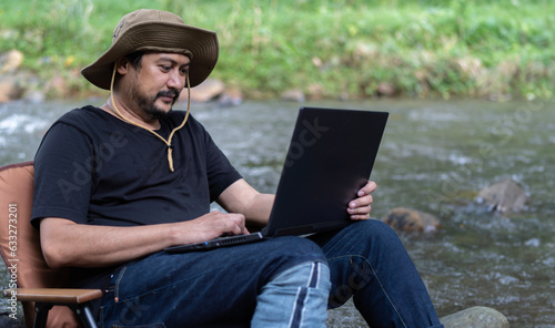 Mature worker Asian man sit down on camping chair and work on the laptop near the stream in the middle of park