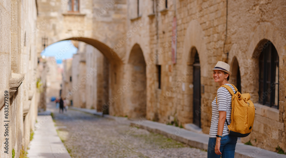 Fototapeta premium summer trip to Rhodes island Greece. Young Asian woman in striped tshirt and hat walks Street of Knights of Fortifications castle. female traveler visiting southern Europe. Unesco world heritage site