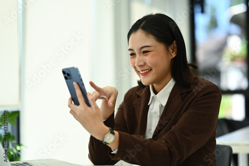 Happy female entrepreneur in luxury suit using smartphone during break from work at office