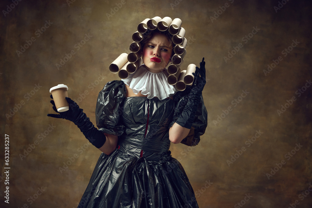 Portrait grimacing woman with paper tube wig in black dress made of garbage bags holding smartphone and coffee cup with gloved hands