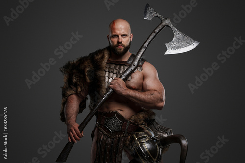 A strong and intimidating Viking man in a beard and bald head, dressed in animal fur and light armor, with a helmet hanging from his belt, brandishing a huge axe against a gray background