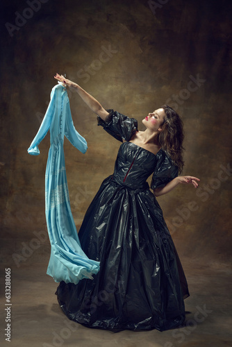 Full body portrait of attractive young woman wearing, black balldress made of garbage bags holding blue midi dress over vintage background.