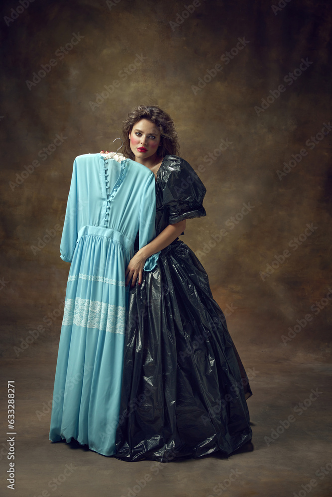 Full body portrait of attractive young woman wearing, black balldress made of garbage bags holding blue midi dress and looking at camera.