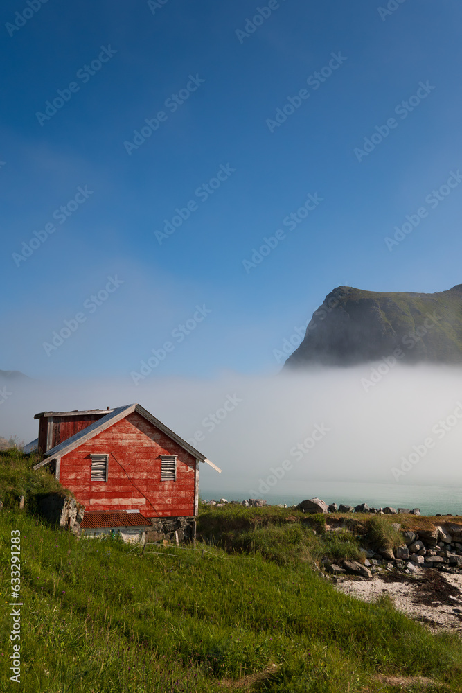 Red norwegian house in a fjord on a bright sunny morning, with fog clouds rolling in from the sea