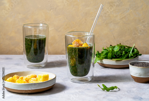 Green smoothie in glasses made with greens, frozen mango, and chia seeds, grey and yellow background, ingredients