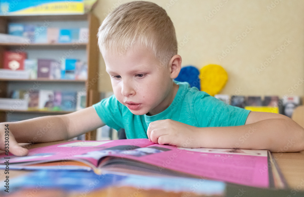 Boy in library. Seven year Ukrainian guy is sitting at a table in library and reading book. Concept of normal learning, thirst for knowledge, desire to learn new things. Blurred background and letters Boy in library. Seven year Ukrainian guy is sitting at a table in library and reading book. Concept of normal learning, thirst for knowledge, desire to learn new things. Blurred background and letters