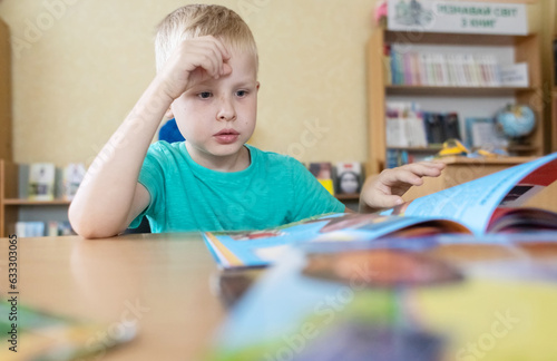 Boy in library. Seven year Ukrainian guy is sitting at a table in library and reading book. Concept of normal learning, thirst for knowledge, desire to learn new things. Blurred background and letters