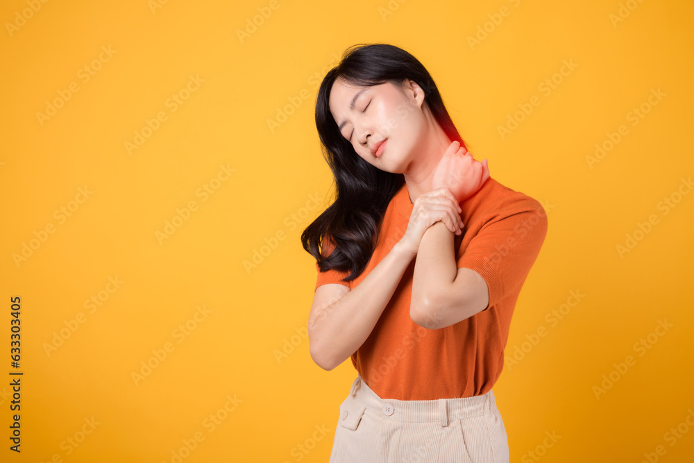 Concerned Asian woman in her 30s, wearing an orange shirt, holds her painful neck on yellow background. office syndrome health care concept. Concerned Asian woman in her 30s, wearing an orange shirt, holds her painful neck on yellow background. office syndrome health care concept.