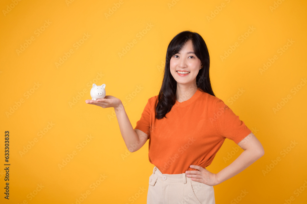 Radiant Asian woman in her 30s holding piggy bank, hands on hips, on vibrant yellow background. Financial growth concept.