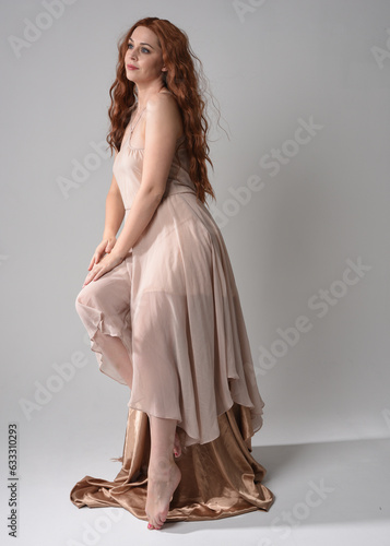 Full length portrait of beautiful female model with long brunette hair wearing a creamy pink gown dress. graceful sitting pose isolated on white studio background.