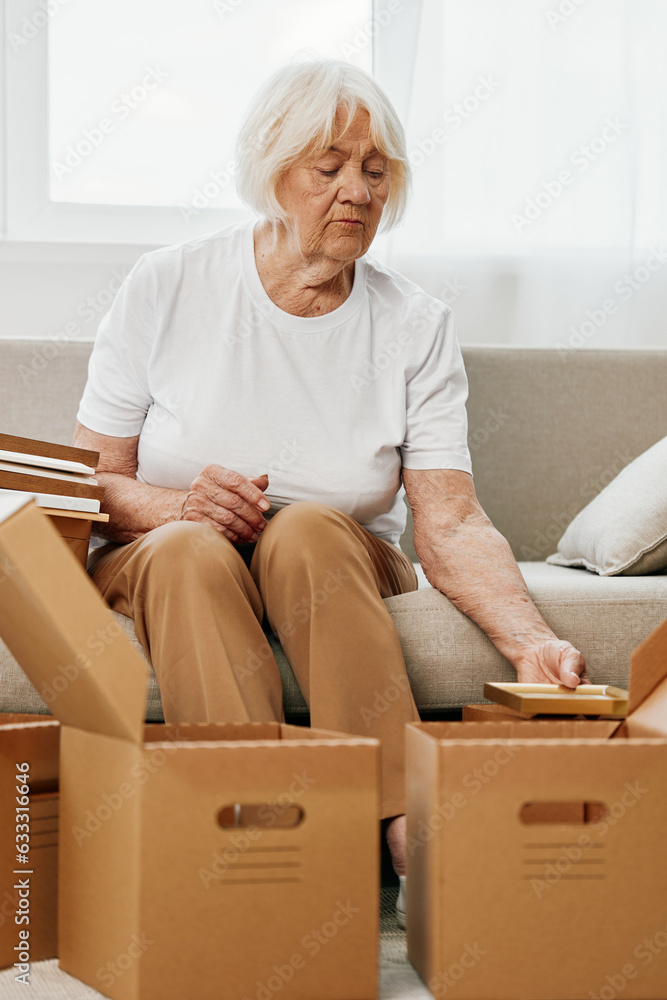 elderly woman sits on a sofa at home with boxes. collecting things with memories albums with photos and photo frames moving to a new place cleaning things and a happy smile. Lifestyle retirement. elderly woman sits on a sofa at home with boxes. collecting things with memories albums with photos and photo frames moving to a new place cleaning things and a happy smile. Lifestyle retirement.
