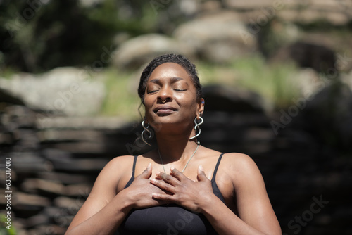 A self-described nonbinary, queer person clasps their hands against their chest and looks up towards their sun with their eyes closed. They express a moment of spiritual joy mixed with serene calm.
