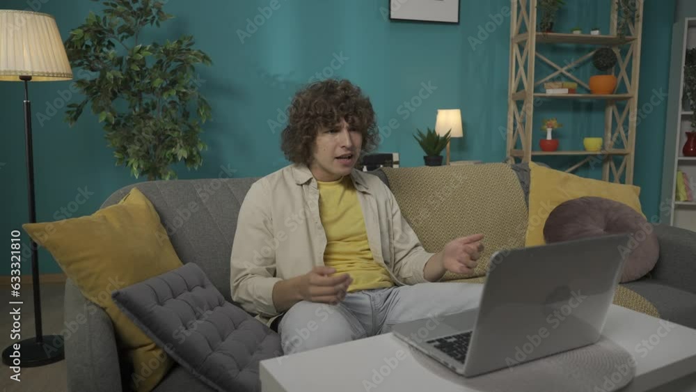 Young man in casual clothing, sitting on the couch in the living room and talking by a video call using a laptop HDR BT2020 HLG Material. Young man in casual clothing, sitting on the couch in the living room and talking by a video call using a laptop HDR BT2020 HLG Material.