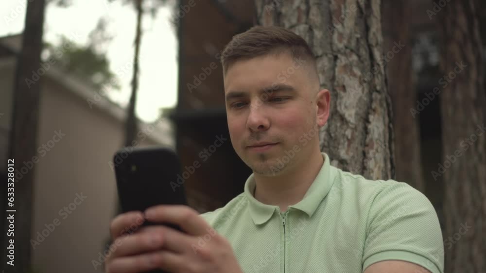 A young man sits under a tree near a country house with a phone in his hands. A man is texting on a smartphone in nature on the grass. Closeup. A young man sits under a tree near a country house with a phone in his hands. A man is texting on a smartphone in nature on the grass. Closeup.