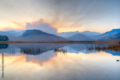 SMOKE FROM A WINTER GRASSFIRE reflects in a drakensberg mountain lake