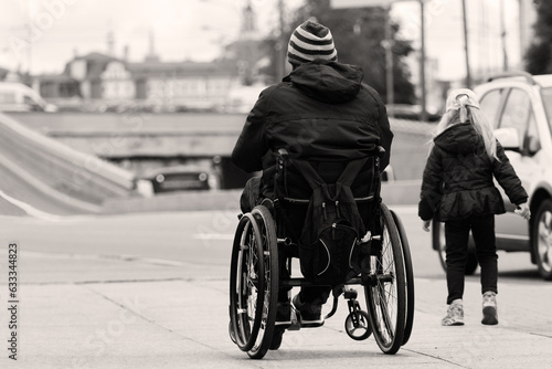 Obraz na płótnie A disabled man rides in a wheelchair along the sidewalk, an elementary school daughter runs nearby, black and white photo