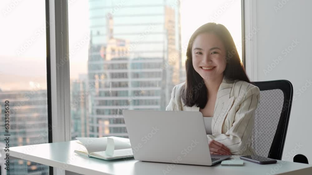 Young pretty smiling Asian business woman employee executive sitting in corporate office with laptop. Happy professional businesswoman looking at camera sitting at desk. Portrait Young pretty smiling Asian business woman employee executive sitting in corporate office with laptop. Happy professional businesswoman looking at camera sitting at desk. Portrait