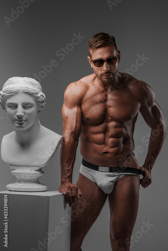 Attractive muscular male model wearing sunglasses and underwear stands confidently next to ancient Greek bust on a gray background