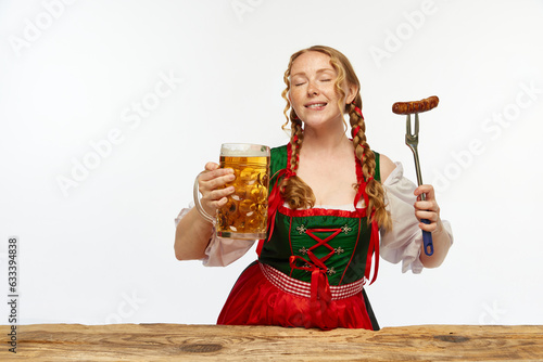 Dreaming redhead woman dressed traditional dirndl, serves appetizing Bavarian sausage on fork and huge beer chole. Concept of Oktoberfest. photo