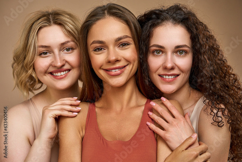 Slika na platnu Close-up portrait of three beautiful different young women on a beige background who are smiling