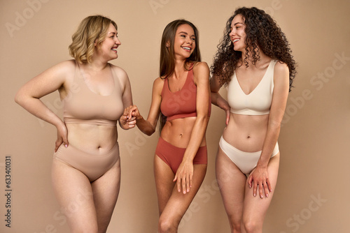 Three beautiful adult women with different types of figures and looks wearing comfortable sports underwear standing on a beige background and laughing.Enjoying and accepting their bodies.