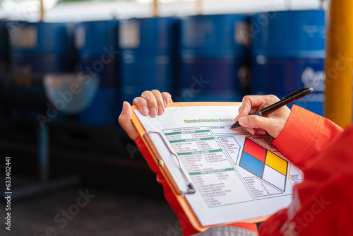 A safety officer is checking on the hazardous material checklist form with chemical storage area at the factory as background. Industrial safety working scene, selective focus. photo