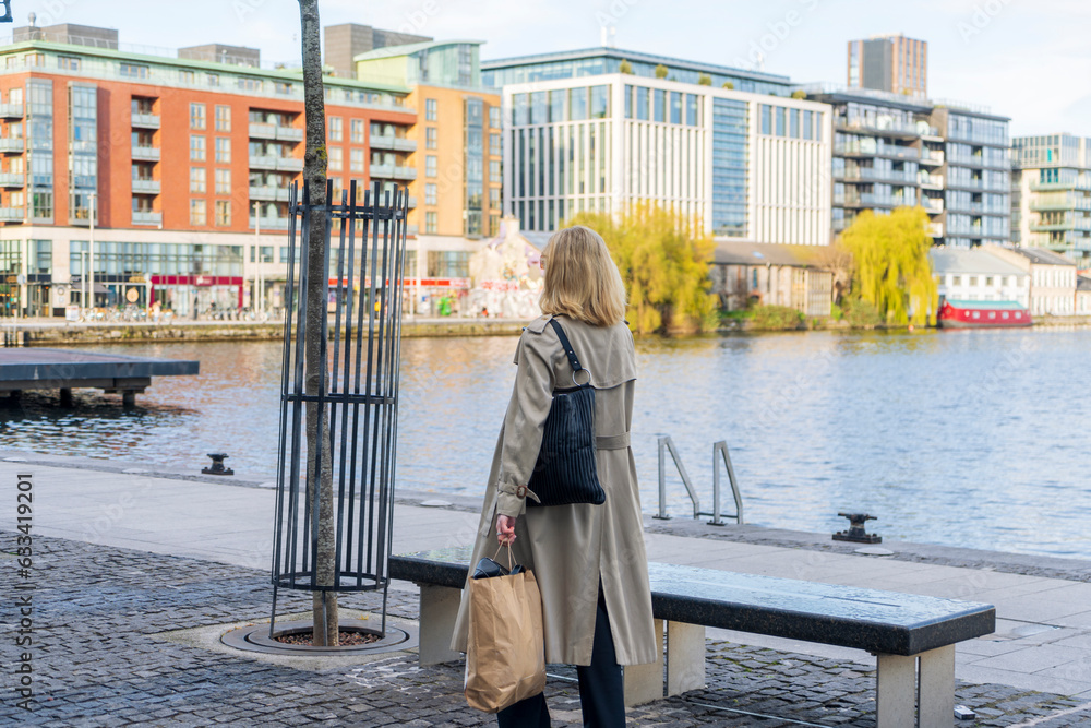 Obraz premium Blonde woman wearing a long beige coat and sunglasses walks after shopping with a paper bag and a bag in Grand Canal Square Dublin, Docklands, Dublin, Ireland