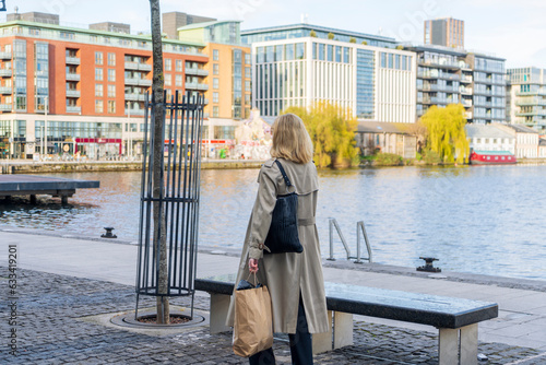 Wallpaper Mural Blonde woman wearing a long beige coat and sunglasses walks after shopping with a paper bag and a bag in Grand Canal Square Dublin, Docklands, Dublin, Ireland Torontodigital.ca