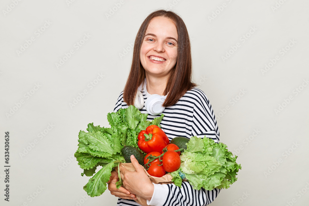 Grocery shopping. Enjoying green diet. Fresh greenery and vegetables. Smiling woman wearing striped casual shirt carrying vegetable isolated over gray background Grocery shopping. Enjoying green diet. Fresh greenery and vegetables. Smiling woman wearing striped casual shirt carrying vegetable isolated over gray background