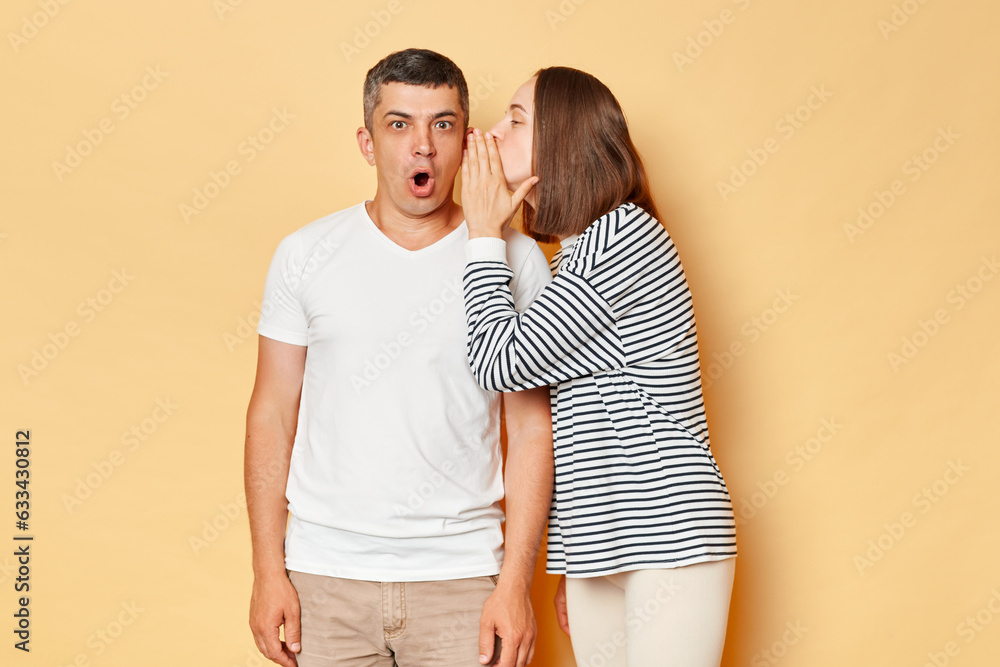 Shocked man hearing gossips from woman talkign to his ear posing with widely opened mouth being amazed and susrprised wearing casual clothing standing isolated over beige background Shocked man hearing gossips from woman talkign to his ear posing with widely opened mouth being amazed and susrprised wearing casual clothing standing isolated over beige background