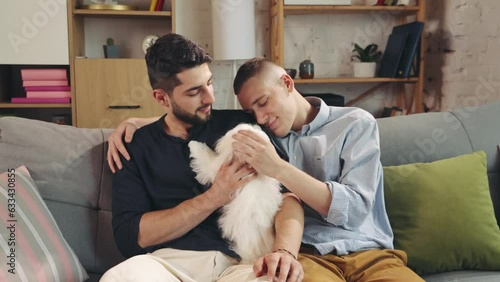 Cute, loving homosexual couple, handsome young men sitting on couch in living room at home and hugging with dog. Showing feelings. Concept of relationship, happiness, animals, lgbt community photo