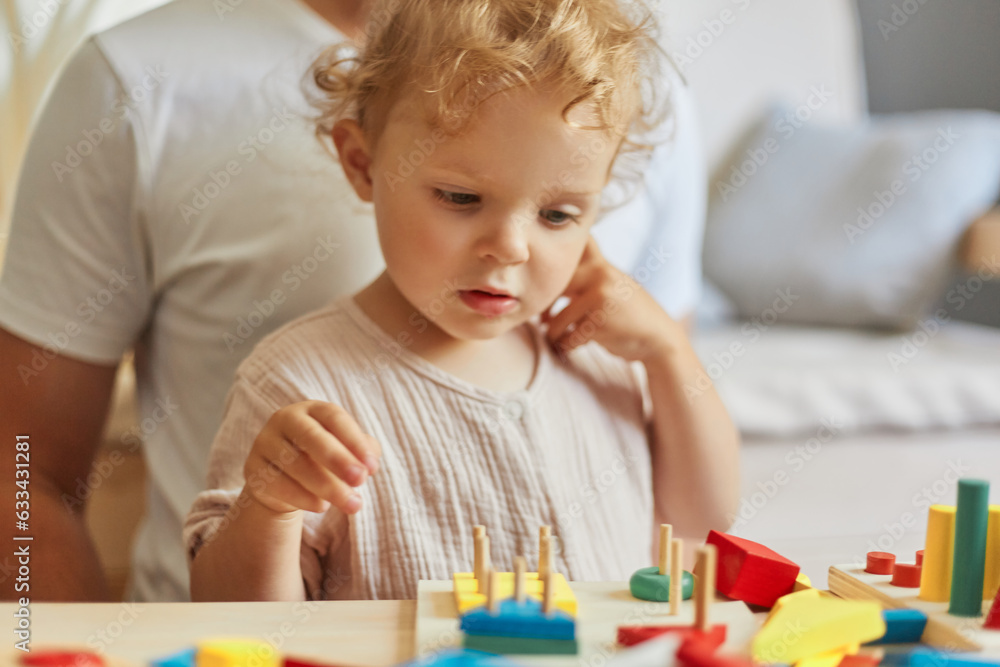 Creative child's playful learning. Beautiful wooden toys for fun. Kindergarten room activities. Curious concentrated wavy haired blonde toddler baby playing wooden educational toys at home interior. Creative child's playful learning. Beautiful wooden toys for fun. Kindergarten room activities. Curious concentrated wavy haired blonde toddler baby playing wooden educational toys at home interior.