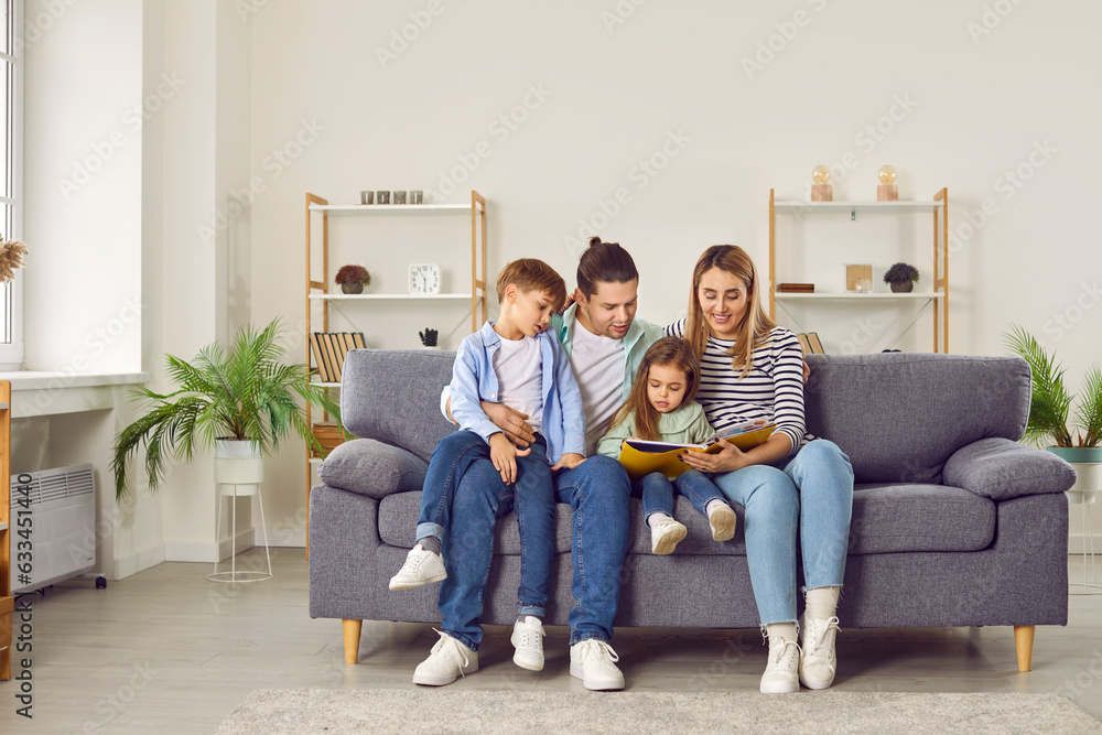 Full length photo of young happy family couple reading book with two kids boy and girl sitting on the sofa at home and smiling. Parents spending time with children. Leisure time together concept. Full length photo of young happy family couple reading book with two kids boy and girl sitting on the sofa at home and smiling. Parents spending time with children. Leisure time together concept.