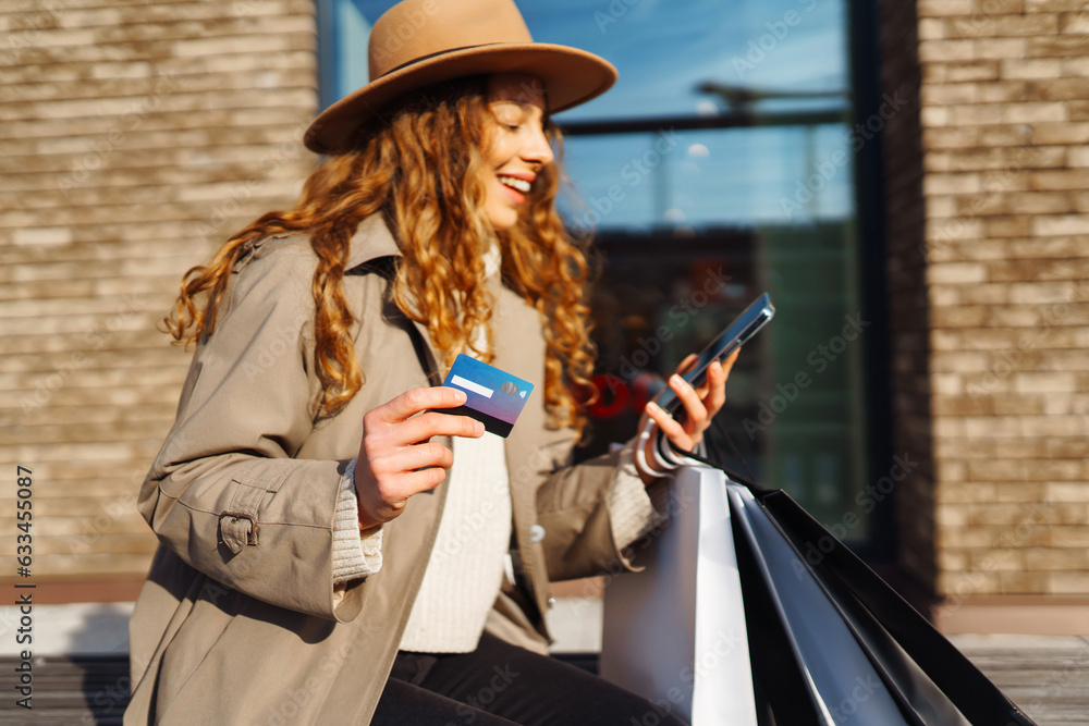 Beautiful woman in hat with shopping bags and holding credit card. Curly woman shopping with credit card, walking on sunny street. E-commerce, internet banking. Lifestyle concept.