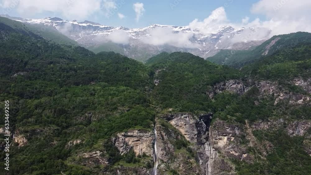 Aerial drone view of Alpine mountain peaks with snow in clouds in summer, Italian Alps, Piedmont, Italy. Rocks covered with forest and waterfall called Cascata di Novalesa flowing down in foreground Aerial drone view of Alpine mountain peaks with snow in clouds in summer, Italian Alps, Piedmont, Italy. Rocks covered with forest and waterfall called Cascata di Novalesa flowing down in foreground