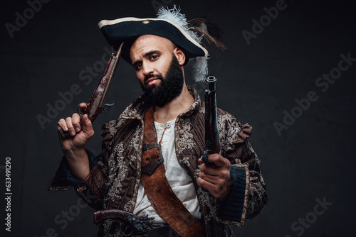 Attractive pirate with a black beard, wearing a vest and hat, holding two muskets against a dark textured wall