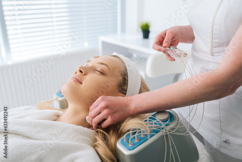 Close-up side view of smiling young woman patient preparing for myostimulation procedure. Beautician putting electrodes of myostimulator during bioresonance therapy in medical clinic. photo