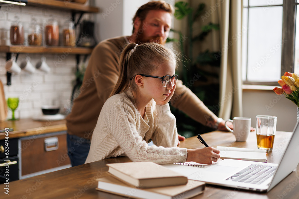 Preadolescent kid in eyeglasses writing on notebook near laptop, books and blurred father at home Preadolescent kid in eyeglasses writing on notebook near laptop, books and blurred father at home