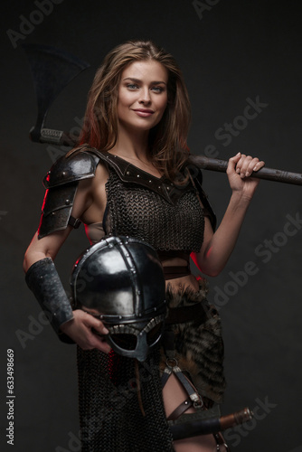 Beautiful Viking girl with a cheerful smile dressed in a chainmail top and fur skirt posing with a two-handed axe illuminated by red backlight against a textured gray wall