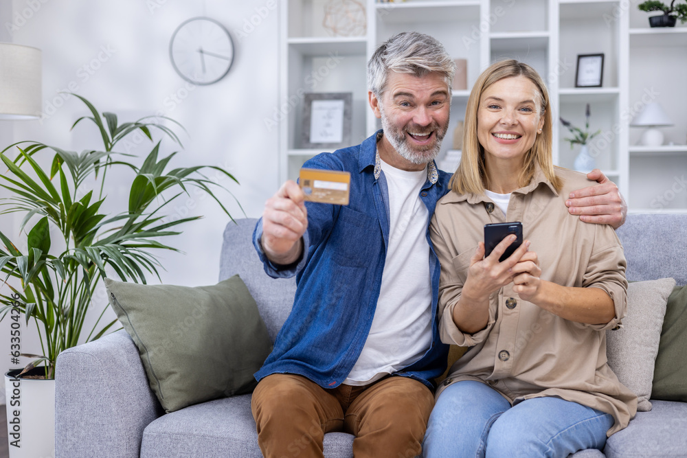 Portrait of mature married couple husband and wife woman sitting on sofa in living room and looking at camera, satisfied online shoppers of online store, holding bank credit card and phone Portrait of mature married couple husband and wife woman sitting on sofa in living room and looking at camera, satisfied online shoppers of online store, holding bank credit card and phone