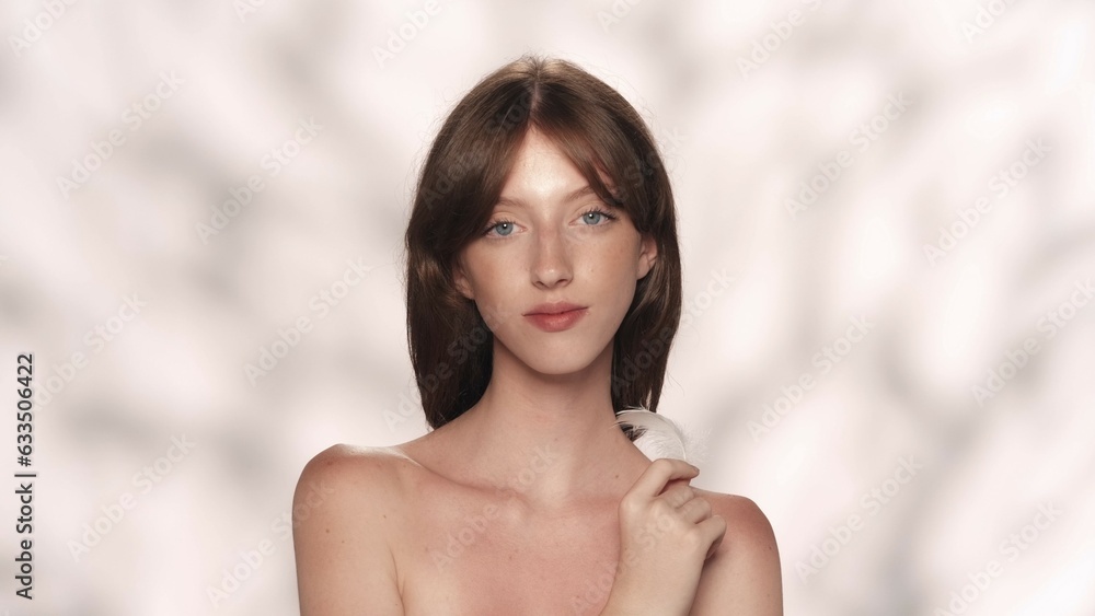 Attractive model with long sliky hair and soft makeup touching her skin with feather, looking at the camera Attractive model with long sliky hair and soft makeup touching her skin with feather, looking at the camera