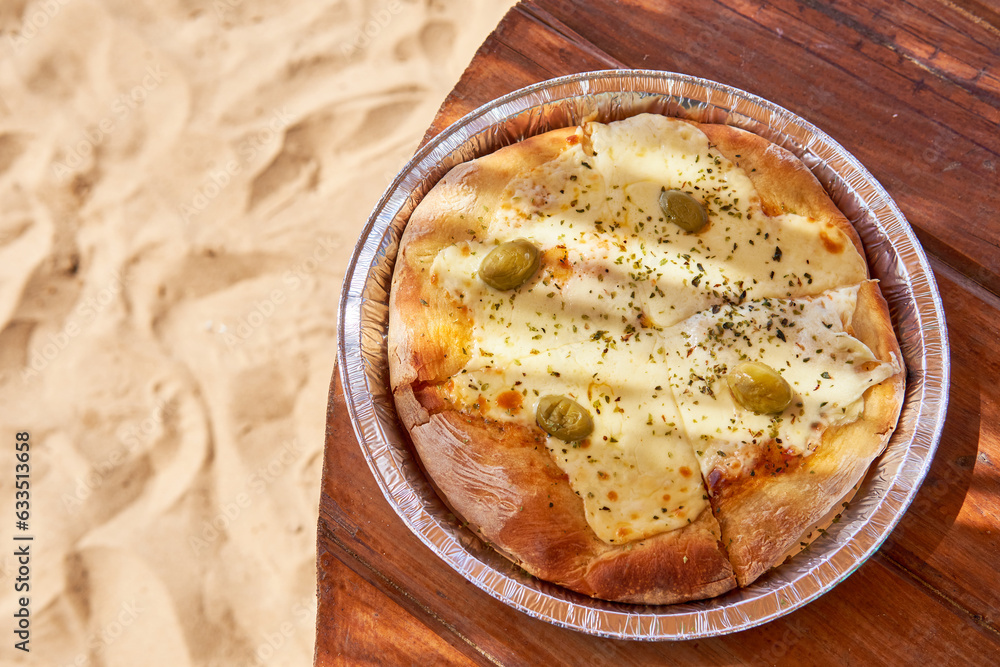 pizza with mozzarella and olives on an aluminum tray on a wooden table on the sand. Fast food to enjoy a summer day at the beach. Copy space pizza with mozzarella and olives on an aluminum tray on a wooden table on the sand. Fast food to enjoy a summer day at the beach. Copy space