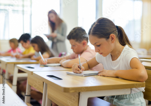 Schoolchildren sitting at desks in classroom. Female teacher staning beside desks. © JackF