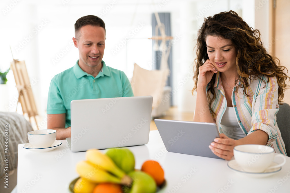 Cheerful young couple planning budget over laptop at table. Young man and woman calculating monthly expenses at home. They are in casuals drinking coffee. Cheerful young couple planning budget over laptop at table. Young man and woman calculating monthly expenses at home. They are in casuals drinking coffee.