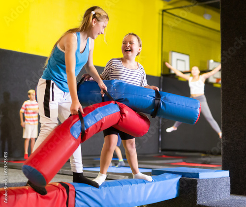 Expressive tween girl fighting by soft pugil stick with her female friend while standing on inflatable beam over pit with foam cubes on bouncy playground