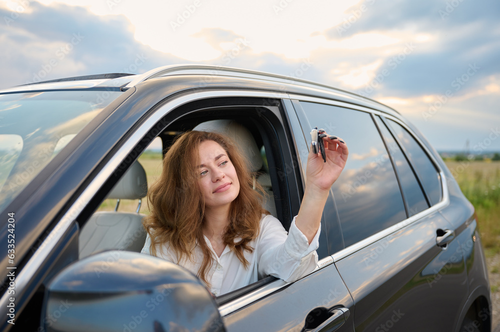 Yung beautiful happy girl holds the keys to a new car in her hand. Close up view of happy smiling business girl with red hair sitting inside the car. Woman posing inside car with key.
