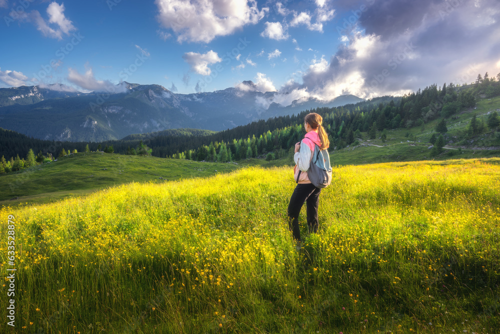 Girl on the hill with yellow flowers and green grass in beautiful alpine mountain valley at sunset in summer. Landscape with young woman in alps, rees, sky with clouds. Travel and Hiking. Slovenia
