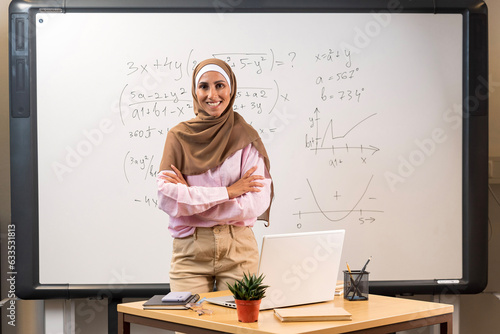 Education concept. Young beautiful Arabic teacher in a hijab stands in the classroom in front of the blackboard with her arms crossed. Confident happy tutor ready to start school year and lesson.  photo