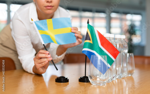 Unrecognizable woman preparing room for international negotiations and communication discussions of leaders. Lady sets miniatures flags of South Africa and Sweden on table. Unfocused shot